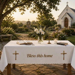 May include: A white rectangular tablecloth featuring a scalloped edge, embroidered with two gold crosses and the text "Bless this home" in a centered script font. The fabric is draped over a table, with an open book and a candle placed on top.