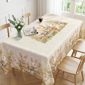 May include: A rectangular Easter-themed tablecloth with a cream background and a repeating pattern of bunnies, flowers, and Easter eggs. The central design features a detailed illustration of bunnies in a basket surrounded by flowers. The table is set with chairs, a vase of white flowers, and a cup of coffee.