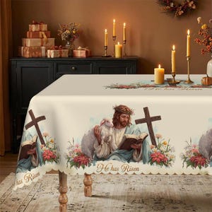 May include: Off-white Easter tablecloth featuring a religious illustration of Jesus with a lamb, a cross, and the text "He has Risen." The scalloped edge is decorated with flowers. Candles and gifts are in the background.