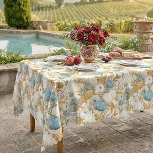 Può includere: Una tovaglia rettangolare con un motivo ripetuto di zucche e fogliame bianco, blu e dorato. Un vaso in ceramica riempito di rose rosse si trova al centro, circondato da piatti bianchi, una ciotola di frutti rossi e pane affettato su un tagliere.