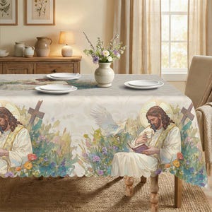 May include: A beige tablecloth with a religious illustration featuring Jesus, a lamb, a cross, and a dove. The design includes colorful flowers and a scalloped edge. The table is set with white plates.