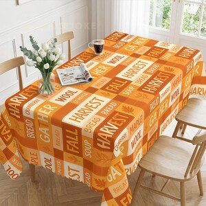Può includere: Una tovaglia rettangolare con un motivo a quadri nei toni dell'arancione e della crema, decorata con parole a tema autunnale tra cui Harvest, Gather, Fall, Bread, Soup, Maple, Cranberry e Wool, insieme a piccole illustrazioni di foglie.