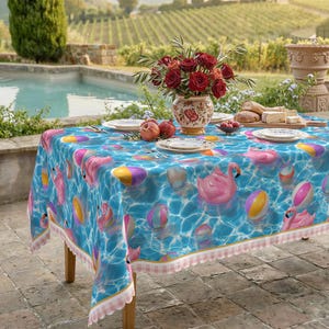 May include: A rectangular tablecloth with a blue water pattern featuring pink flamingos, beach balls, and inner tubes. The table is set with plates, silverware, and a vase of red roses. The tablecloth has a pink and white gingham trim.