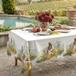 May include: A white rectangular tablecloth featuring a repeating watercolor-style print of a figure in robes sitting in a landscape with sheep. The table is set with white plates, silverware, a floral vase with red roses, and a wooden cutting board.