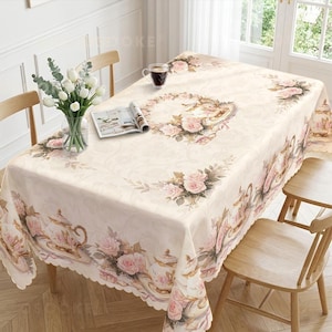 Puede incluir: Un mantel rectangular de color crema claro con un patrón repetitivo de rosas rosadas y teteras de estilo vintage. La tela tiene un borde festoneado y una textura sutilmente grabada, cubriendo una mesa con un jarrón de tulipanes blancos.