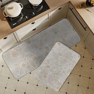 May include: Three kitchen mats with a floral design in shades of beige and white. The mats are rectangular and oval-shaped, with a soft, textured surface. The mats are placed on a tiled floor in front of a kitchen counter.