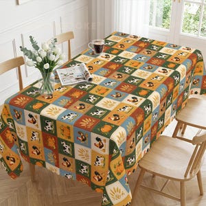 May include: A rectangular tablecloth featuring a repeating grid pattern of squares with illustrations of roosters, cows, pumpkins, and wheat stalks in shades of orange, green, blue, and white, draped over a dining table with a vase of white tulips.