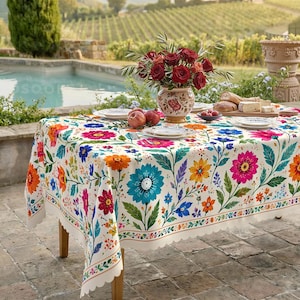 Puede incluir: Un mantel rectangular con base blanca que presenta un vibrante patrón floral multicolor con motivos botánicos en azul, naranja, rosa y verde. Los bordes tienen un acabado festoneado y la tela cubre una mesa de madera.