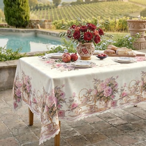 Puede incluir: Un mantel rectangular con fondo crema que presenta un patrón repetitivo de rosas rosadas, teteras y tazas de té. La mesa está puesta con platos blancos, un jarrón de cerámica lleno de rosas rojas y una tabla de madera con pan.