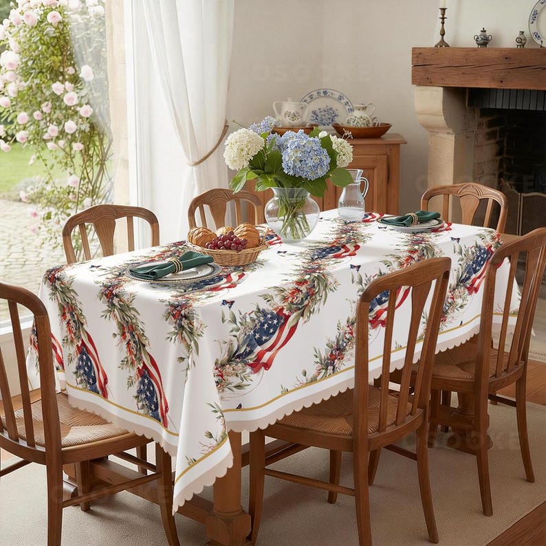 Pu&ograve; includere: Una tovaglia rettangolare bianca con un motivo ripetuto di bandiere americane drappeggiate intrecciate con ghirlande di foglie verdi e piccole bacche rosse. La tovaglia ha un bordo smerlato ed &egrave; stesa su un tavolo da pranzo in legno.