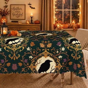Op de afbeelding: Een donkergroen tafelkleed met een herhalend patroon van zwarte raven op takken binnen cirkelvormige kaders, omringd door sierlijk goudkleurig krulwerk en herfstkleurige bladeren in tinten oranje, bruin en paars.