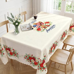 May include: A rectangular cream-coloured tablecloth featuring a scalloped edge and a floral border of red roses and white daisies. The centre displays a graphic of a wooden cross draped with an American flag, surrounded by flowers and the text God Bless America.