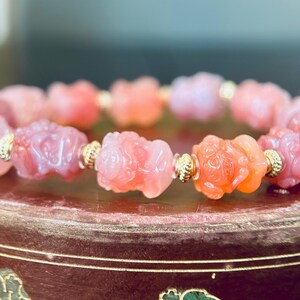 May include: A stretch bracelet featuring carved pink stone beads shaped like small creatures, interspersed with small, ribbed gold-tone metal spacer beads. The beads exhibit varying shades of translucent pink and coral, arranged in a circular pattern.