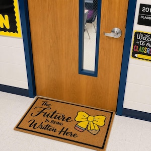 May include: A rectangular tan coir doormat with a black border and the black text The Future Is Being Written Here. The design includes a yellow bow graphic with a pencil tip at the bottom, positioned in front of a wooden classroom door.