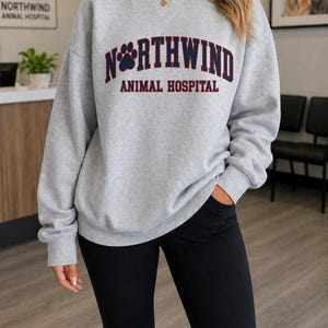 May include: A light grey sweatshirt featuring the text "NORTHWIND ANIMAL HOSPITAL" in navy and red. The sweatshirt has a crew neck and long sleeves. The image also shows black trousers.