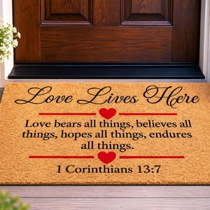 May include: A rectangular tan coir doormat featuring the black text Love Lives Here, followed by the verse Love bears all things, believes all things, hopes all things, endures all things, and 1 Corinthians 13:7, accented by two red hearts.