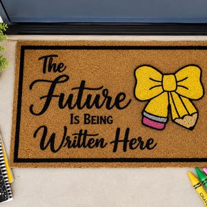 May include: A rectangular tan coir doormat with a black border and the black text The Future Is Being Written Here. To the right of the text is a yellow bow graphic made of two pencils with pink erasers and tan tips. The mat has a coarse texture.