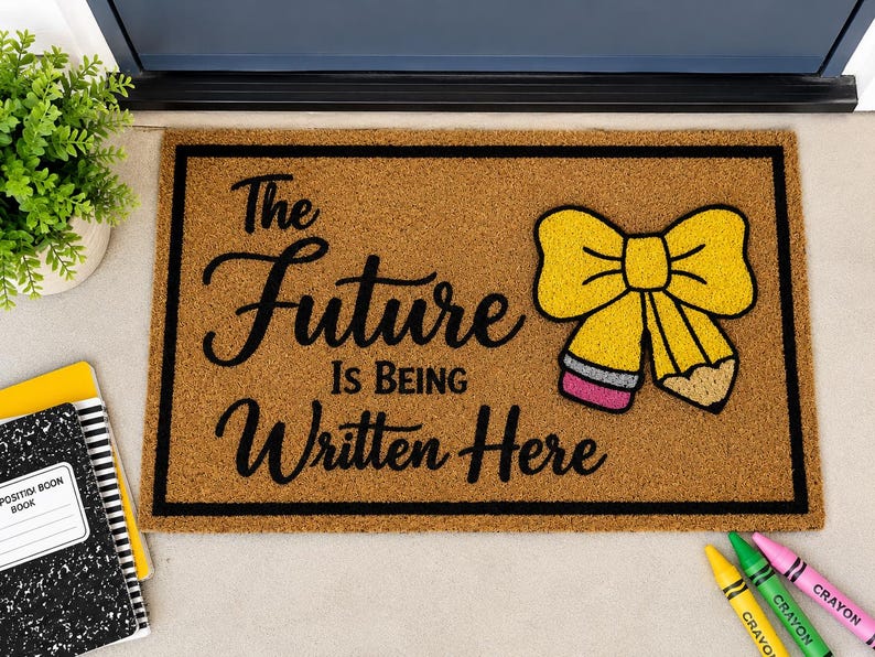 May include: A rectangular tan coir doormat with a black border and the black text The Future Is Being Written Here. To the right of the text is a yellow bow graphic made of two pencils with pink erasers and tan tips. The mat has a coarse texture.