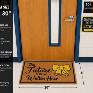 May include: A rectangular tan doormat measuring 45.72 cm by 76.2 cm with a black border. The mat features the black text 'The Future Is Being Written Here' and a graphic of a yellow bow tied around a pencil with a pink eraser and grey lead.