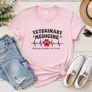 Może przedstawiać: Jasnoróżowy t-shirt z krótkim rękawem z czarnym napisem "VETERINARY MEDICINE" nad czarną linią tętna z czerwonym odciskiem łapy pośrodku oraz czarnym napisem "Because People Are Gross" wydrukowanym pod grafiką.