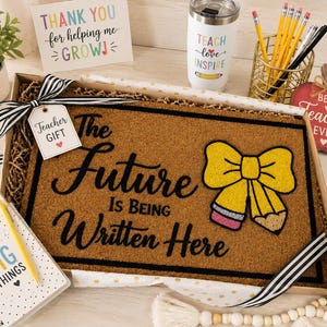 May include: A rectangular brown coir doormat in a gift box featuring the black text The Future Is Being Written Here and a yellow pencil-shaped bow graphic. The mat is decorated with a black and white striped ribbon and a Teacher Gift tag.