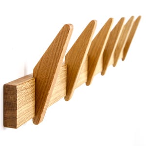 Peut inclure: Porte-manteau mural en bois avec une série de crochets triangulaires inclinés. Le porte-manteau est en bois clair et est fixé à un mur blanc. Le design est moderne et minimaliste.