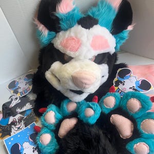 May include: A black, white, and blue furry fursuit head with matching paw pads and stickers. The head has pink accents and a white muzzle. The paw pads are black with blue and pink details. Several stickers with cartoon animal designs are also visible.