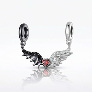 May include: A silver charm with a heart-shaped red gem in the center, flanked by a black bat wing on one side and a white, gem-studded angel wing on the other. The charm has two silver loops for attachment.