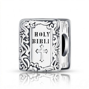 Puede incluir: Un charm plateado con forma de libro, con las palabras "HOLY BIBLI" y una cruz en la portada. El charm tiene un borde adornado y está ligeramente abierto, revelando las páginas. El charm es probablemente para una pulsera o collar.