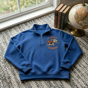 May include: A blue quarter-zip sweatshirt with the text "MY DOG IS A SUPERDOG" and a cartoon dog graphic. The dog is wearing a cape and flying. The sweatshirt has a ribbed collar and cuffs.