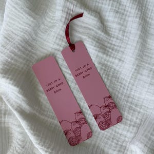 Könnte beinhalten: Zwei rechteckige rosafarbene Lesezeichen mit einer dunkelroten Strichzeichnung von Erdbeeren und Himbeeren am unteren Rand. Jedes Lesezeichen zeigt den Text "LOST IN A BERRY GOOD BOOK" in dunkelroter Schrift. Ein Lesezeichen hat ein dunkelrotes Band.