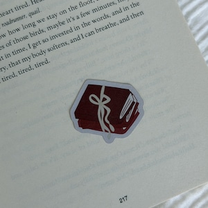 Könnte beinhalten: Ein gestanzter Aufkleber mit einem Stapel aus zwei dunkelroten Büchern, die mit einem hellgrauen Band zusammengebunden sind. Der Aufkleber liegt auf einer offenen Buchseite mit sichtbarem schwarzem Text und der Seitenzahl 217 unten.