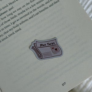 Könnte beinhalten: Ein gestanzter Aufkleber in Form einer gefalteten Zeitung mit dem in Schwarz gedruckten Text Plot Twist. Der Aufkleber zeigt eine kleine Illustration eines Herzens und eines Sterns auf der Titelseite. Er liegt auf der Seite eines offenen Buches.