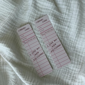 Könnte beinhalten: Zwei rechteckige Lesezeichen im Design alter Bibliothekskarten. Jedes Lesezeichen ist weiß mit rosa horizontalen Linien und dem Text LIBRARY CARD, TITLE, AUTHOR, DUE DATE, BROWSERS NAME sowie I LIKE MY MEN FICTIONAL in dunkelroter Schrift.