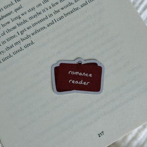 Könnte beinhalten: Ein rechteckiger Aufkleber mit abgerundeten Ecken, einem dunkelroten Hintergrund und dem weißen Text "romance reader" in einer lockeren, handschriftlichen Schriftart. Der Aufkleber befindet sich auf einer Buchseite mit sichtbarem Text.
