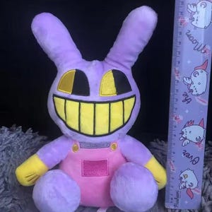 Puede incluir: Un peluche de conejo en tonos morado, amarillo y rosa. El conejo tiene una gran sonrisa con dientes amarillos, ojos negros y detalles amarillos. Lleva un peto rosa. Una regla con un dise&ntilde;o de dibujos animados est&aacute; al lado del conejo.