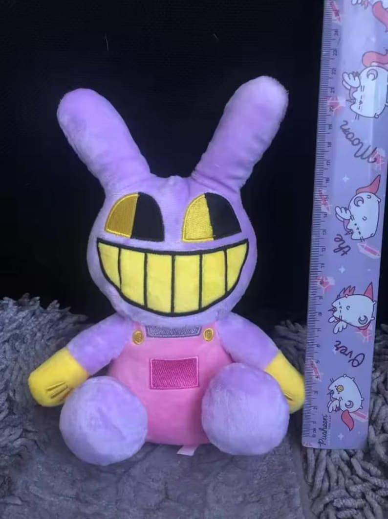 Puede incluir: Un peluche de conejo en tonos morado, amarillo y rosa. El conejo tiene una gran sonrisa con dientes amarillos, ojos negros y detalles amarillos. Lleva un peto rosa. Una regla con un dise&ntilde;o de dibujos animados est&aacute; al lado del conejo.