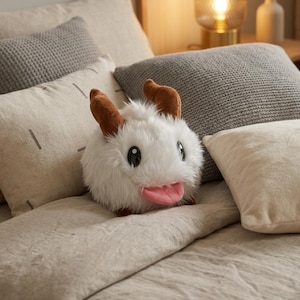 May include: A plush white creature with brown horns and a pink tongue, resting on a bed with gray and cream-colored pillows. The stuffed animal has large black eyes and a fluffy texture, creating a whimsical appearance.