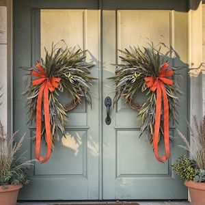 May include: Two matching teardrop-shaped wreaths made of long, slender green leaves and dried grasses. Each wreath features a prominent, bright orange fabric bow with long, trailing ribbons hanging down the center of the foliage arrangement.