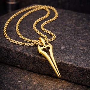 Può includere: Una collana con pendente dorato con un design allungato e stilizzato e un elemento a forma di cuore nella parte superiore. Il pendente è appeso a una catena dorata abbinata. La collana è appoggiata su una superficie scura e strutturata, mettendo in risalto i dettagli del gioiello.