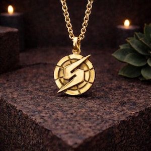 Puede incluir: Collar con colgante dorado con un colgante circular con un diseño de rayo estilizado. El colgante cuelga de una cadena dorada a juego. El collar se muestra sobre una superficie texturizada de color marrón oscuro.