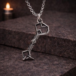 May include: A silver-colored pendant necklace featuring a key-shaped charm. The charm has a geometric design with a keyhole shape at the top and intricate patterns along the stem. The necklace chain is also silver, and the background is a dark, textured surface.