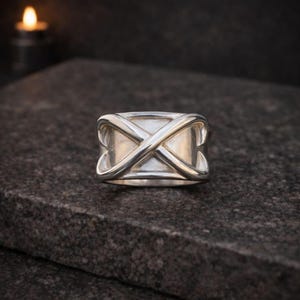 Könnte beinhalten: Ein silberner Ring mit einem X-förmigen Design auf der Vorderseite. Der Ring ist breit und hat eine polierte Oberfläche. Der Ring liegt auf einer dunkelgrauen Oberfläche.