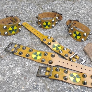 Puede incluir: Cinco pulseras de cuero color amarillo mostaza con tachuelas de metal plateado y un s&iacute;mbolo circular de peligro de radiaci&oacute;n en amarillo y negro. Cada pulsera incluye un cierre de hebilla met&aacute;lica y varios orificios de ajuste.