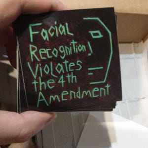 Può includere: Una pila di adesivi quadrati con la scritta "Facial Recognition Violates the 4th Amendment" in verde e il contorno verde di un volto su sfondo scuro. Gli adesivi sono tenuti da una mano.