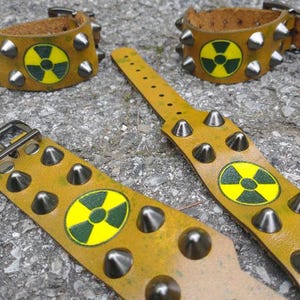 Puede incluir: Tres pulseras de cuero color amarillo mostaza con tachuelas c&oacute;nicas de metal plateado y un s&iacute;mbolo circular de peligro de radiaci&oacute;n en amarillo y verde oscuro impreso en el centro. Cada pulsera incluye un cierre de hebilla de metal.
