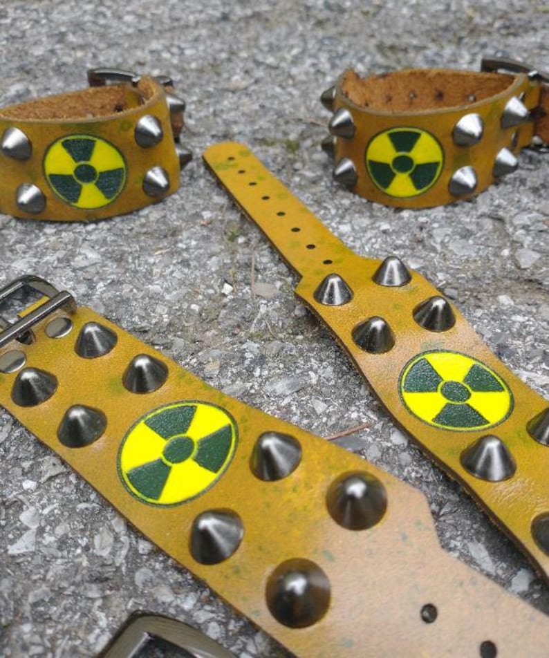 Puede incluir: Tres pulseras de cuero color amarillo mostaza con tachuelas c&oacute;nicas de metal plateado y un s&iacute;mbolo circular de peligro de radiaci&oacute;n en amarillo y verde oscuro impreso en el centro. Cada pulsera incluye un cierre de hebilla de metal.