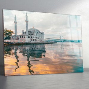 May include: A scenic print of a mosque with two tall minarets and a domed roof, reflected in the water. A bridge is visible in the background under a cloudy sky. The water reflects the colors of the sunset.