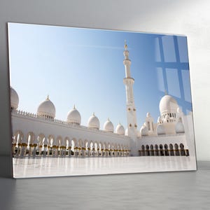 Op de afbeelding: Een ingelijste print van een witte moskee met meerdere koepels en een hoge minaret tegen een helderblauwe lucht. De moskee heeft gewelfde loopbruggen en gouden accenten, die reflecteren op een gepolijst wit oppervlak.