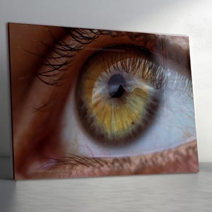 Puede incluir: Primer plano de un ojo humano, mostrando detalles intrincados. El iris es una mezcla de tonos amarillos y marrones, con una pupila oscura en el centro. Largas pestañas enmarcan el ojo, y la piel circundante es de color marrón rojizo cálido. La imagen es una impresión.
